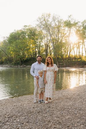 Family of three posing by a sunlit river with lush green trees in the background during golden hour.