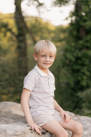 A young blonde boy in a striped polo shirt and shorts sits on a rock outdoors, smiling at the camera.
