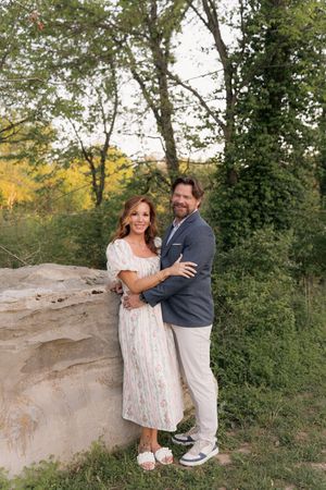 A couple embraces outdoors near a stone wall, surrounded by lush green trees, woman in floral dress, man in blazer.