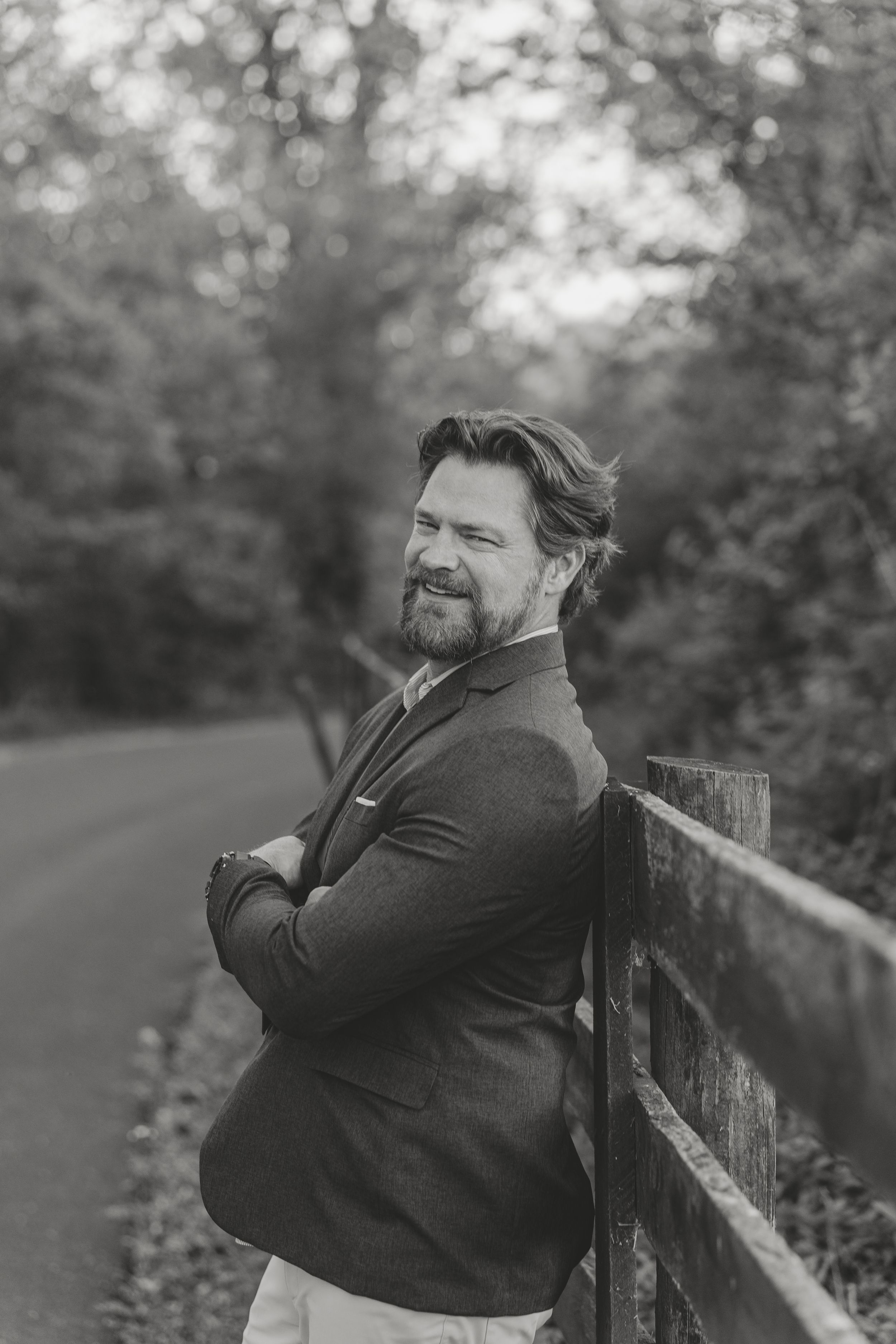 Black and white portrait of a bearded man in a blazer leaning on a fence along a tree-lined road.