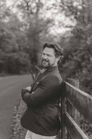 Black and white portrait of a bearded man in a blazer leaning on a fence along a tree-lined road.