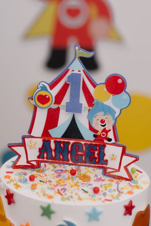 Circus-themed first birthday cake topper for Angel featuring a big top tent, clown, balloons, and colorful sprinkles.
