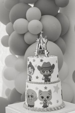 Black and white two-tier birthday cake with cartoon characters, cowboy cat, stars, and Angel topper, surrounded by balloons.