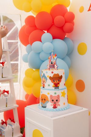 Two-tier birthday cake with cartoon characters, topped with a number 1, surrounded by colorful red, blue, and yellow balloons.