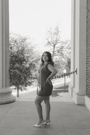 Black and white photo of a young woman in a short dress and heels posing between columns of a classic building.