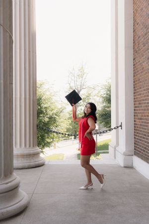 Young woman in red dress holding graduation cap between white columns at university.