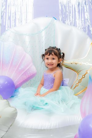 A little girl in a purple and teal mermaid dress sits in a giant shell chair surrounded by pastel balloons.