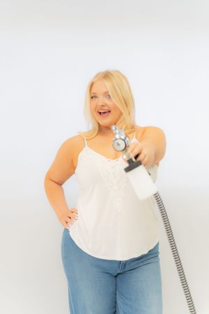 Smiling blonde plus-size woman in white cami and jeans holding a spray tan gun on white background.
