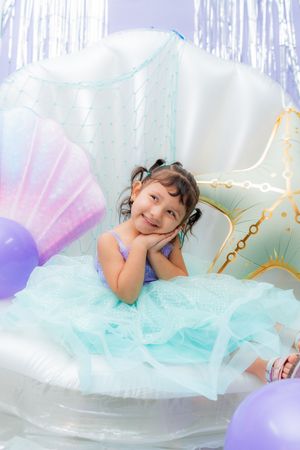 Young girl in teal tutu dress sitting in giant seashell balloon with purple balloons at mermaid birthday party.