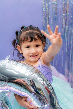 A smiling little girl in a purple tutu holds silver balloons and shows three fingers at a birthday party.