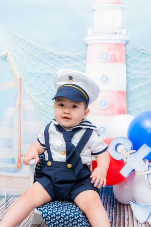 Adorable baby in sailor costume with cap sits before lighthouse backdrop with colorful balloons.