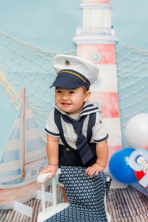 Smiling baby in sailor costume with captain hat poses in nautical themed photo backdrop with lighthouse and sailboat.