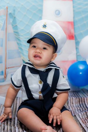 Adorable baby boy in sailor costume and captain hat sitting in nautical themed photo shoot with balloons.