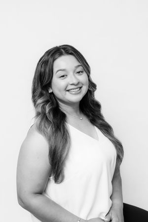 Smiling young woman with long wavy hair wearing a white sleeveless top, posing for a professional headshot photo.