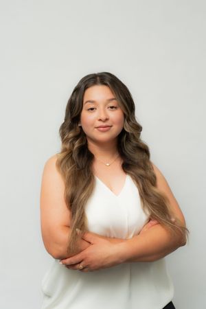 Young woman with long wavy hair in white sleeveless top, arms crossed, smiling against light gray background.