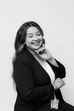 Smiling young woman in black blazer poses confidently for professional headshot in black and white photo.