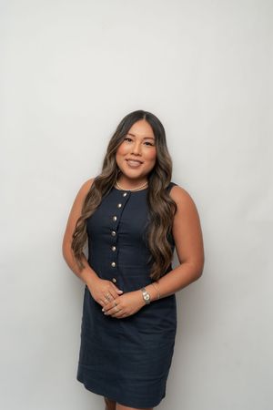 Woman in navy sleeveless button-front dress smiling confidently against a white background.