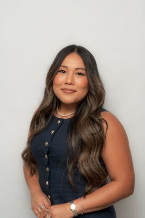 Professional headshot of a smiling young woman with long wavy brown hair wearing a navy sleeveless dress and pearl necklace.