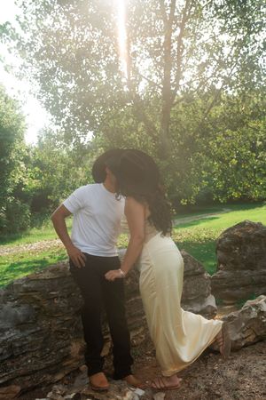 Couple in romantic outdoor setting, woman in elegant gown and man in white tee, standing by rocks in sunlit park.