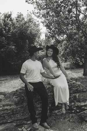 Black and white photo of a couple sitting on a stone wall surrounded by trees, smiling at each other.