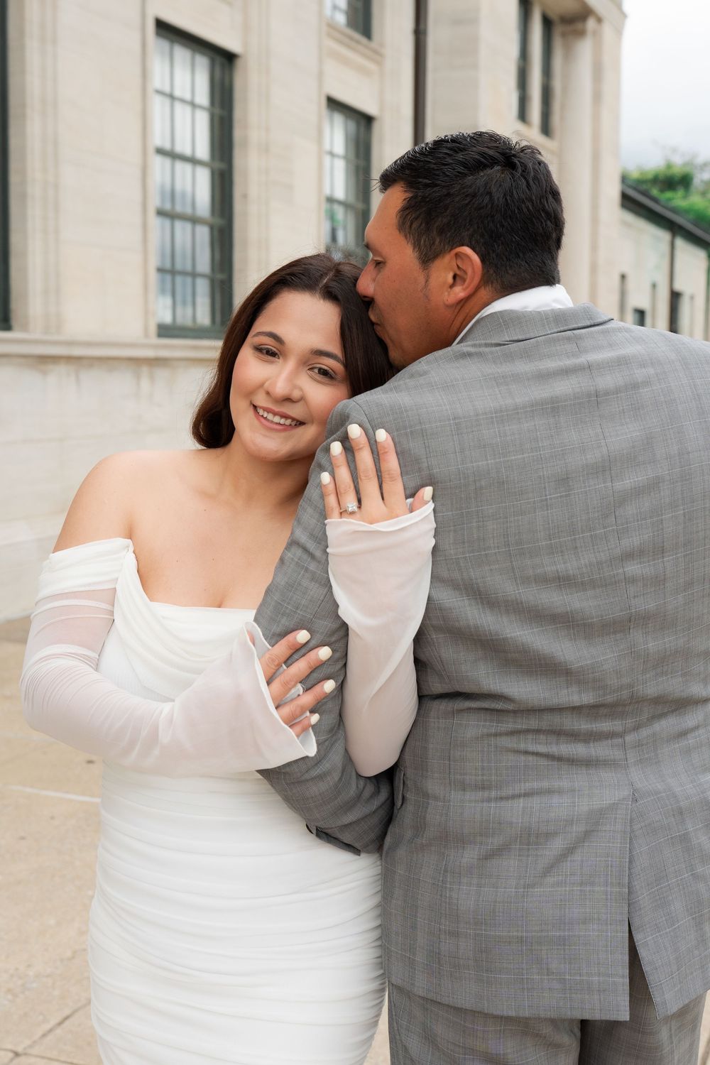 A smiling bride in a white off-shoulder gown with mesh gloves embraces a groom in a gray suit outdoors.