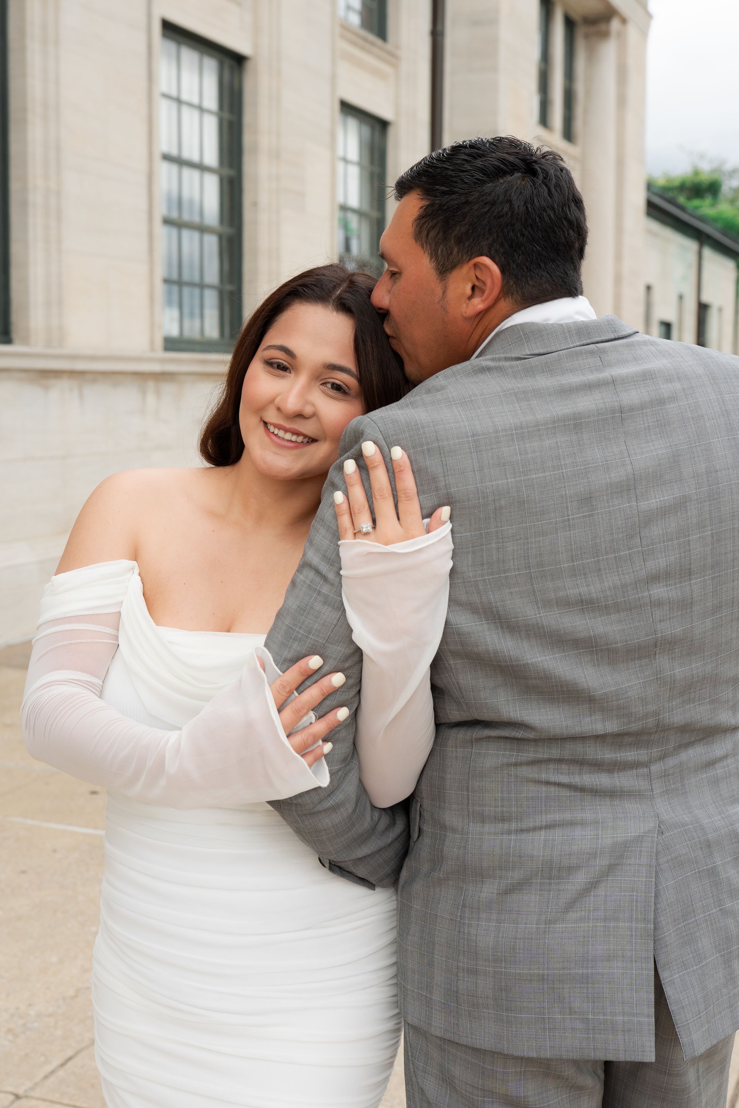 A smiling bride in a white off-shoulder gown with mesh gloves embraces a groom in a gray suit outdoors.