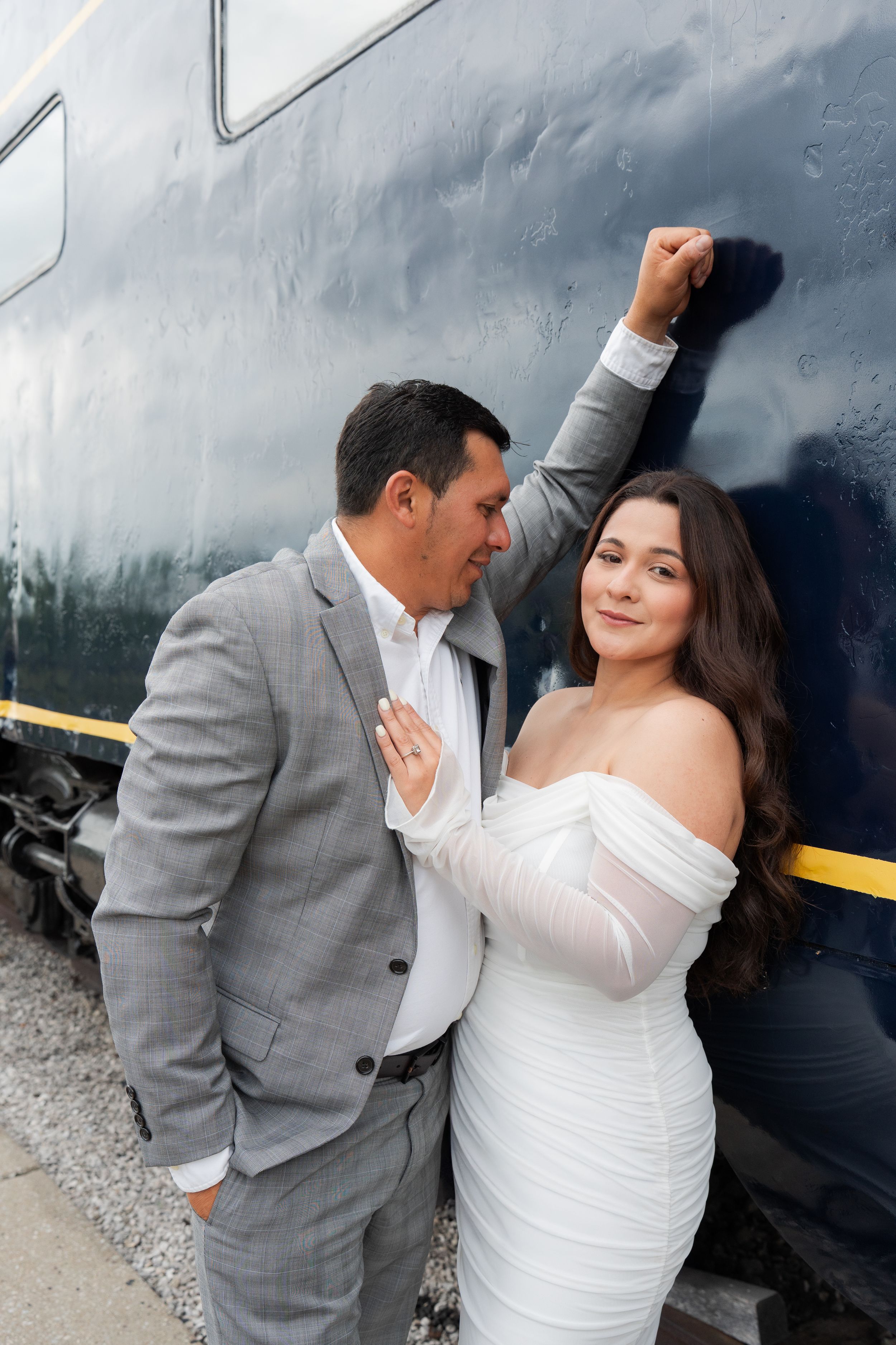 Couple posing romantically by a dark blue train, man in gray suit, woman in white off-shoulder dress.