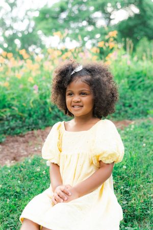 A smiling young girl with curly hair wearing a yellow dress sits outdoors surrounded by green grass and flowers.