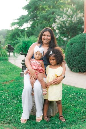 A woman sits outdoors with two young girls in a lush green park, all smiling in colorful summer outfits.