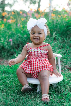 Smiling baby girl in red gingham romper and white bow headband sitting on white chair in garden.