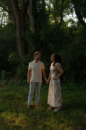A couple holds hands while standing in a lush green forest, the man in a white polo and the woman in a floral dress.