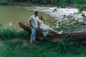 Couple posing by a fallen log near a scenic river surrounded by lush greenery outdoors.