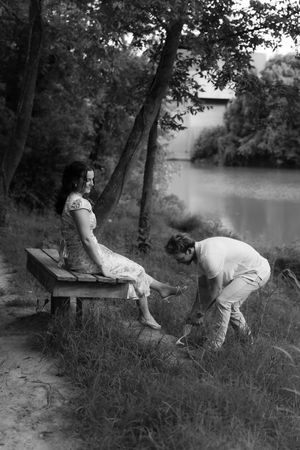 Black and white photo of a couple by a river, woman sitting on bench while man tends to her feet.
