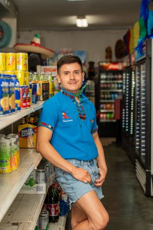 Young man in blue polo shirt posing in a Latin grocery store aisle with beverages and canned goods.