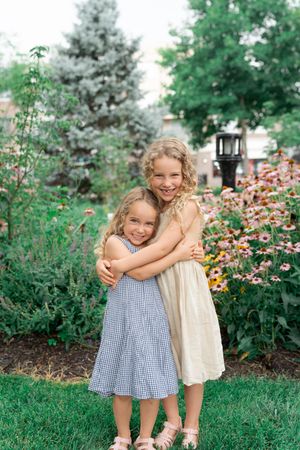Two young girls in dresses hugging and smiling in a lush garden with colorful flowers and greenery.