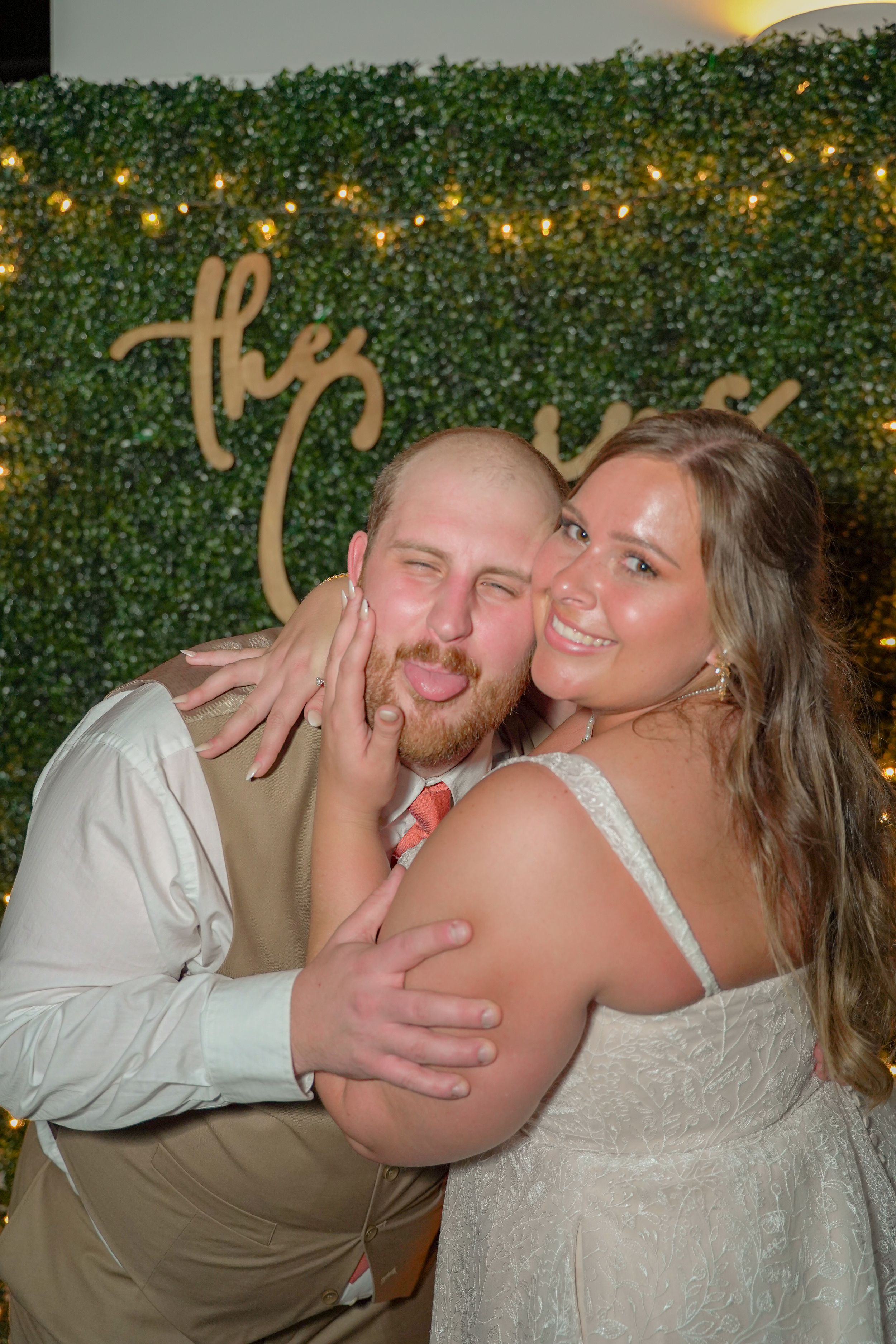 Playful wedding couple posing in front of lit greenery wall.