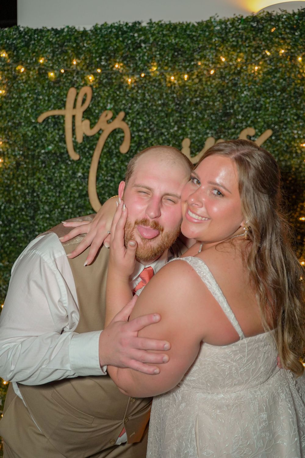 Playful wedding couple posing in front of lit greenery wall.