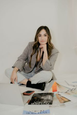 Stylish figure in grey blazer sitting with magazines and phone on white background.