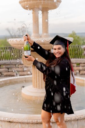 Graduate celebrating with champagne by fountain in traditional cap and gown.