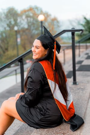 Graduate in cap and gown poses on outdoor steps during sunset celebration.