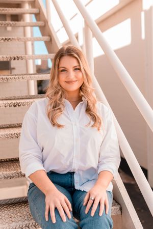 A casual portrait of someone in a white button-down shirt and jeans sitting on carpeted stairs with natural lighting.