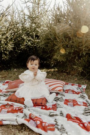 Baby in white dress sitting on plaid blanket outdoors near evergreen trees.