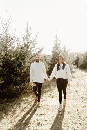Couple holding hands walking through Christmas tree farm on sunny day.