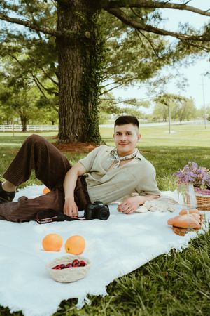 Young man relaxing on white picnic blanket under tree with camera, fruit, bread, and flowers in a park.
