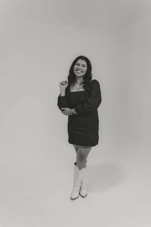 A person in a black dress and white boots poses playfully against a light gray studio backdrop.