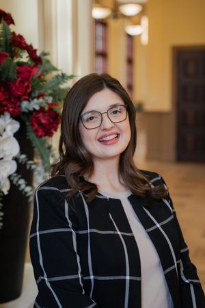 Smiling young woman with glasses wearing a black plaid blazer, posed in an elegant hallway with red floral arrangements.