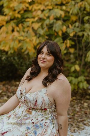 A person in a floral dress poses outdoors against autumn foliage in a natural setting.