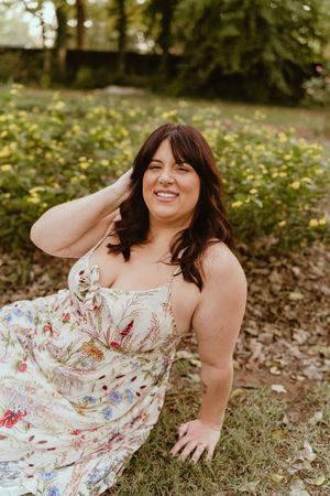 A person in a floral sundress lies on grass in a garden setting during a casual outdoor photoshoot.