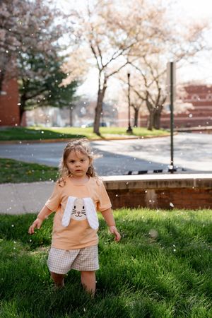 Toddler girl in bunny shirt and plaid shorts standing on green grass with cherry blossom trees in background.