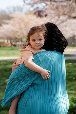 A young girl in a peach dress hugs a woman in a teal outfit outdoors near blooming cherry blossom trees.